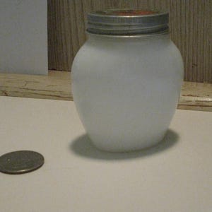 May include: A white, frosted glass jar with a silver lid. A coin rests on the surface next to the jar. The jar has a rounded shape and is likely used for storage or canning.