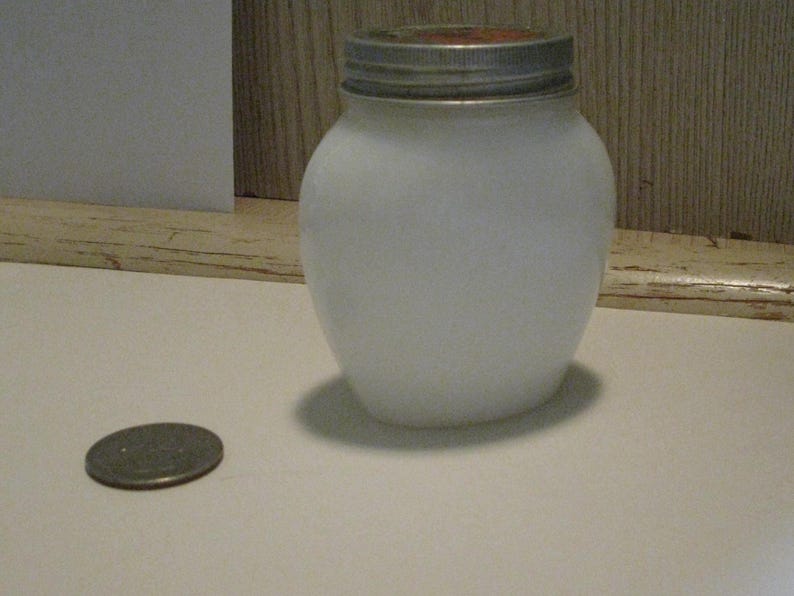 May include: A white, frosted glass jar with a silver lid. A coin rests on the surface next to the jar. The jar has a rounded shape and is likely used for storage or canning.