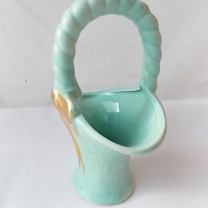 May include: A light blue ceramic basket-shaped vase with a handle. The handle has a twisted rope design. Gold accents are visible on the side of the vase. The vase has a textured, basket-weave pattern on the lower portion.