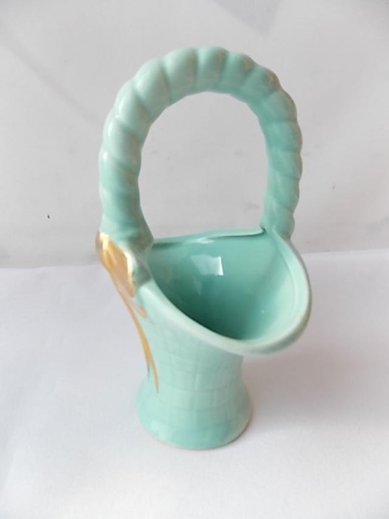 May include: A light blue ceramic basket-shaped vase with a handle. The handle has a twisted rope design. Gold accents are visible on the side of the vase. The vase has a textured, basket-weave pattern on the lower portion.