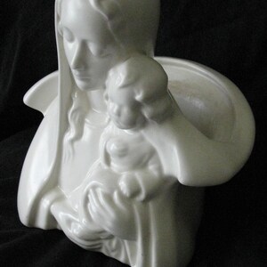 May include: A white ceramic sculpture depicting a woman holding a child. The woman is wearing a draped head covering and holding the child close. The sculpture has a smooth, glossy finish and is set against a dark background.
