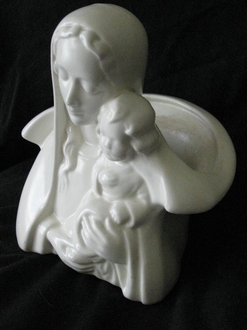 May include: A white ceramic sculpture depicting a woman holding a child. The woman is wearing a draped head covering and holding the child close. The sculpture has a smooth, glossy finish and is set against a dark background.