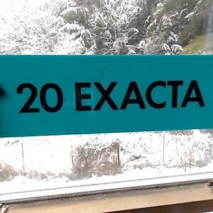 May include: A teal rectangular sign displaying the text "20 EXACTA" in black. The sign is held up in front of a window, with a snowy outdoor scene visible through the glass.