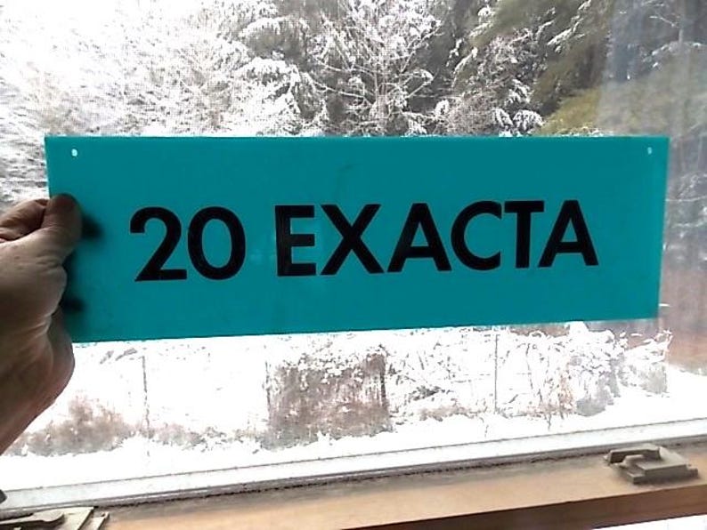 May include: A teal rectangular sign displaying the text "20 EXACTA" in black. The sign is held up in front of a window, with a snowy outdoor scene visible through the glass.