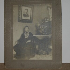 May include: A vintage black and white photograph of a woman seated in a chair, framed within a decorative border. The woman is wearing a dark dress with a lace collar. A framed portrait hangs on the wall behind her.