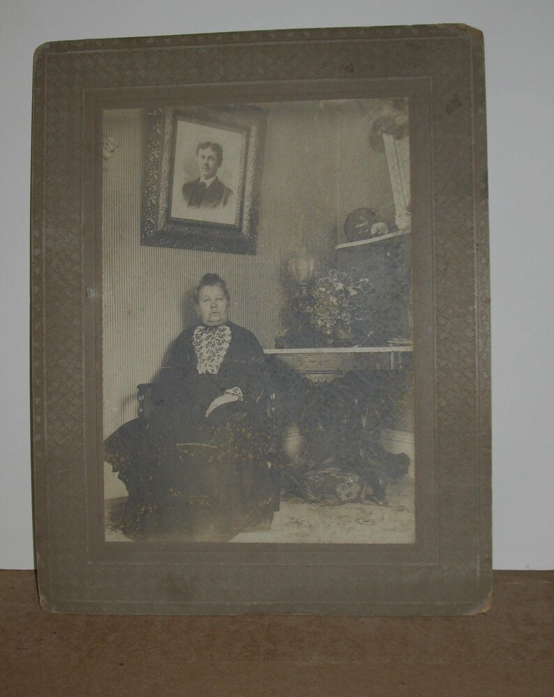 May include: A vintage black and white photograph of a woman seated in a chair, framed within a decorative border. The woman is wearing a dark dress with a lace collar. A framed portrait hangs on the wall behind her.