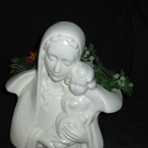 May include: A white ceramic sculpture of a mother and child. The mother is wearing a head covering and holding the child. Green and white decorative foliage is visible behind the sculpture.