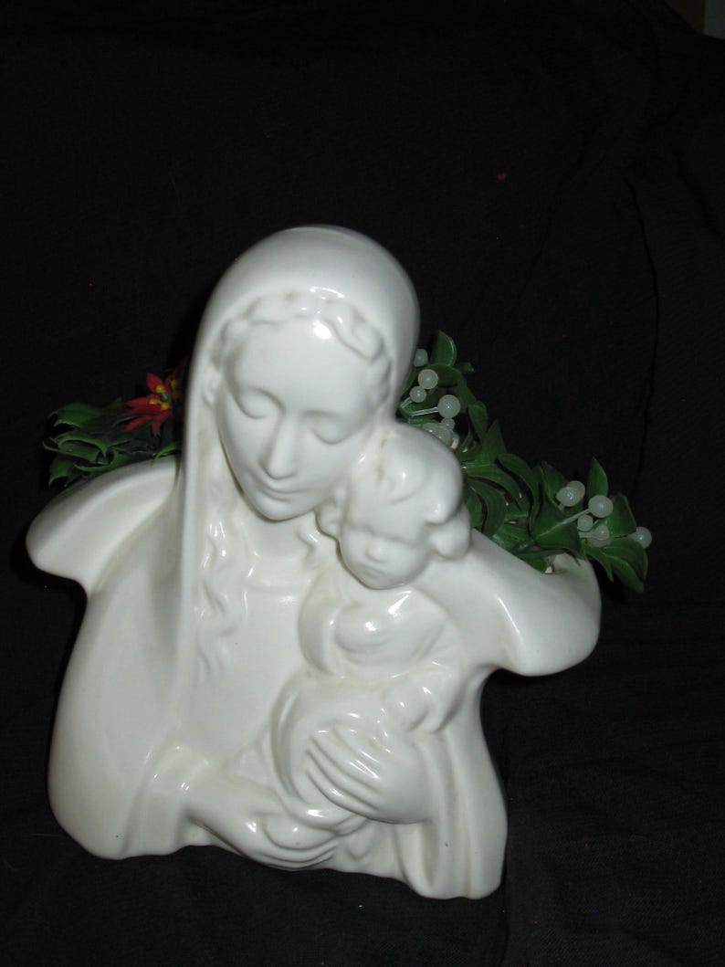 May include: A white ceramic sculpture of a mother and child. The mother is wearing a head covering and holding the child. Green and white decorative foliage is visible behind the sculpture.