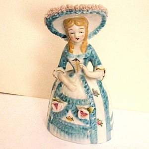 May include: A porcelain figurine of a person wearing a wide-brimmed hat with pink ruffled trim, a blue and white dress with a laced bodice, and floral accents. The figure has blonde hair and is depicted in a standing pose with a bell-shaped skirt.