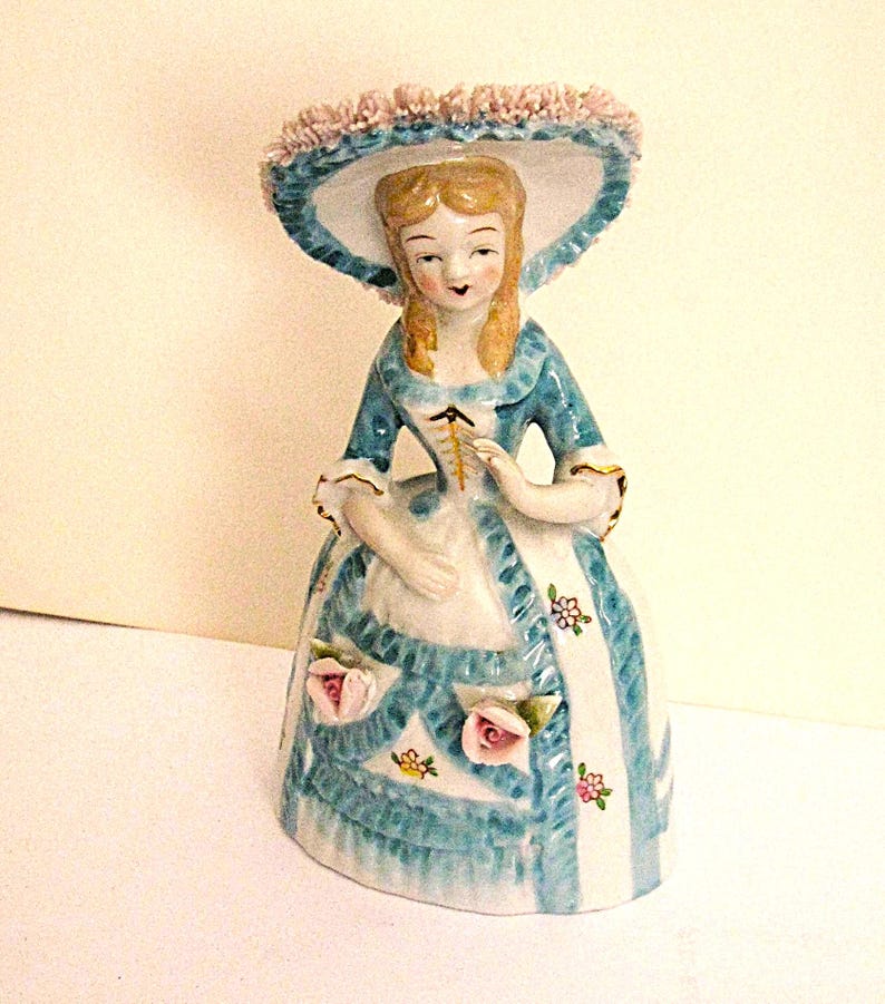 May include: A porcelain figurine of a person wearing a wide-brimmed hat with pink ruffled trim, a blue and white dress with a laced bodice, and floral accents. The figure has blonde hair and is depicted in a standing pose with a bell-shaped skirt.