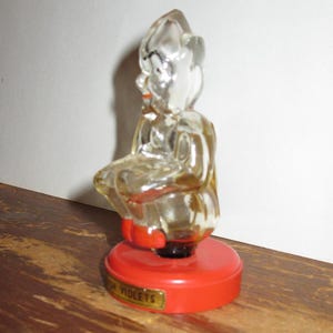 K&ouml;nnte beinhalten: Klare Glasfigur einer sitzenden Figur mit roter Basis. Die Basis hat ein goldenes Etikett mit der Aufschrift "FOR VIOLETS". Die Figur hat einen roten Mund und sitzt auf einer roten, kreisf&ouml;rmigen Basis.