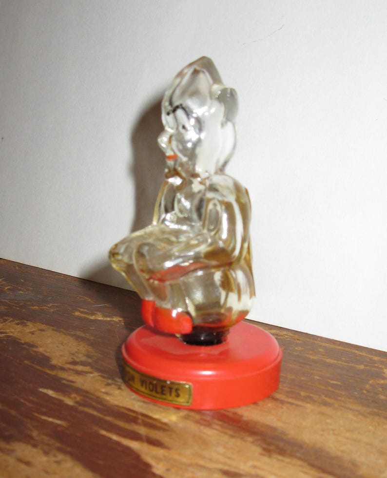 K&ouml;nnte beinhalten: Klare Glasfigur einer sitzenden Figur mit roter Basis. Die Basis hat ein goldenes Etikett mit der Aufschrift "FOR VIOLETS". Die Figur hat einen roten Mund und sitzt auf einer roten, kreisf&ouml;rmigen Basis.