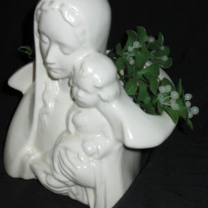 May include: A white ceramic figurine depicting a woman holding a child. The figurine has a smooth, glossy finish and is adorned with a green plant with white berries. The woman is wearing a head covering and has a serene expression.