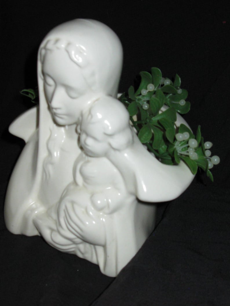May include: A white ceramic figurine depicting a woman holding a child. The figurine has a smooth, glossy finish and is adorned with a green plant with white berries. The woman is wearing a head covering and has a serene expression.