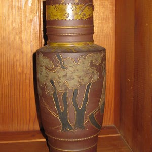 Peut inclure: Un grand pichet cylindrique en c&eacute;ramique brune avec une bande textur&eacute;e jaune moutarde autour du col, orn&eacute;e de petites &eacute;toiles et de points. Le corps pr&eacute;sente un paysage en relief avec des troncs d'arbres noirs et un feuillage vert.