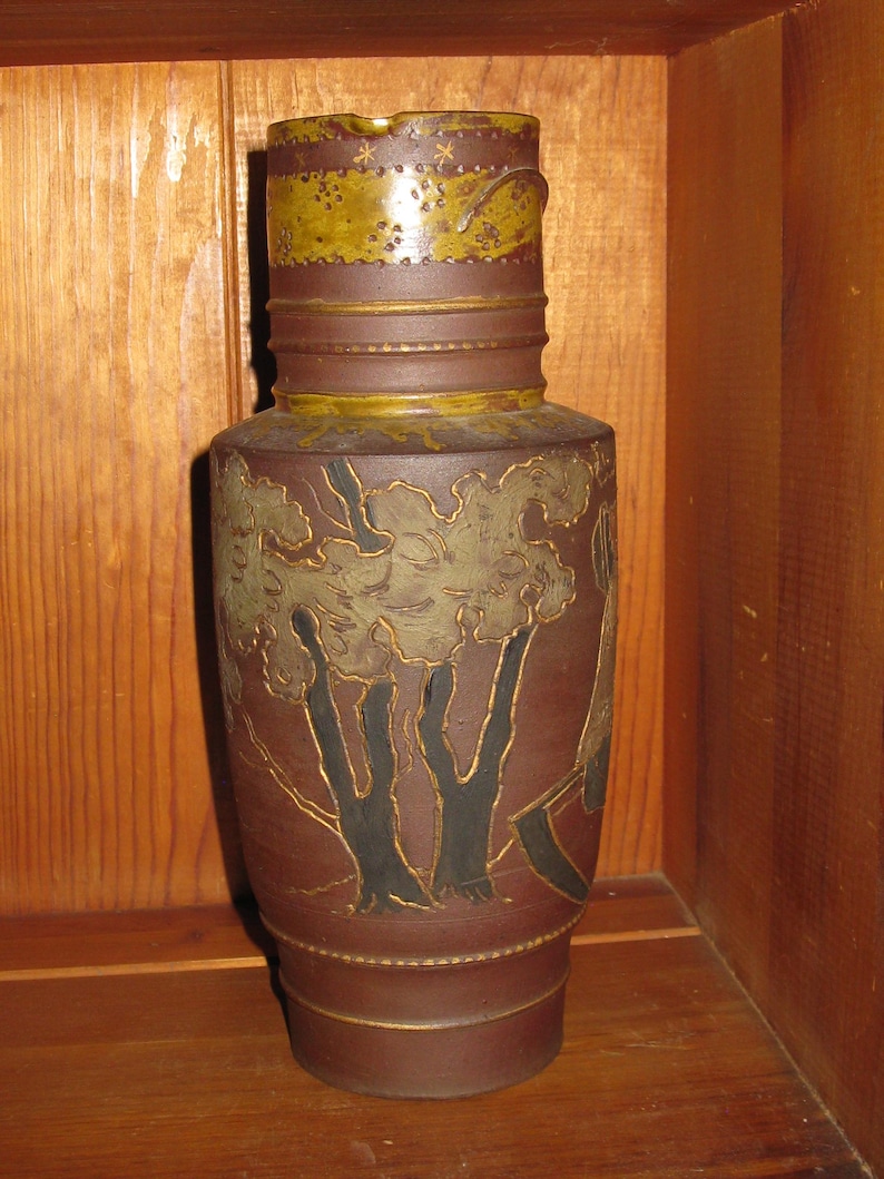 Peut inclure: Un grand pichet cylindrique en c&eacute;ramique brune avec une bande textur&eacute;e jaune moutarde autour du col, orn&eacute;e de petites &eacute;toiles et de points. Le corps pr&eacute;sente un paysage en relief avec des troncs d'arbres noirs et un feuillage vert.