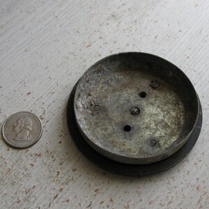 Puede incluir: Un objeto met&aacute;lico circular y desgastado con un borde oscuro, posiblemente una tapa o base. El metal tiene un aspecto desgastado y envejecido con varios agujeros peque&ntilde;os. Una moneda est&aacute; al lado del objeto para la comparaci&oacute;n de tama&ntilde;o.