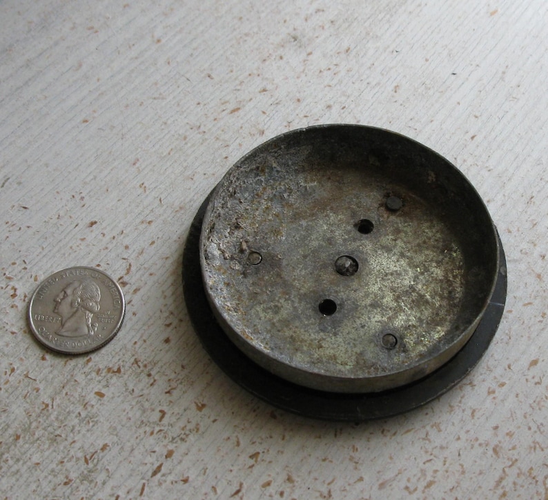 Puede incluir: Un objeto met&aacute;lico circular y desgastado con un borde oscuro, posiblemente una tapa o base. El metal tiene un aspecto desgastado y envejecido con varios agujeros peque&ntilde;os. Una moneda est&aacute; al lado del objeto para la comparaci&oacute;n de tama&ntilde;o.