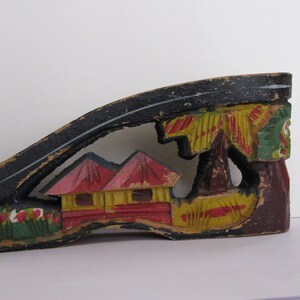 Peut inclure: Un talon de chaussure en bois noir pr&eacute;sentant une d&eacute;coupe peinte &agrave; la main repr&eacute;sentant deux petites maisons aux toits rouges et aux murs jaunes, entour&eacute;es de palmiers verts et de fleurs rouges. La peinture est us&eacute;e et &eacute;caill&eacute;e.