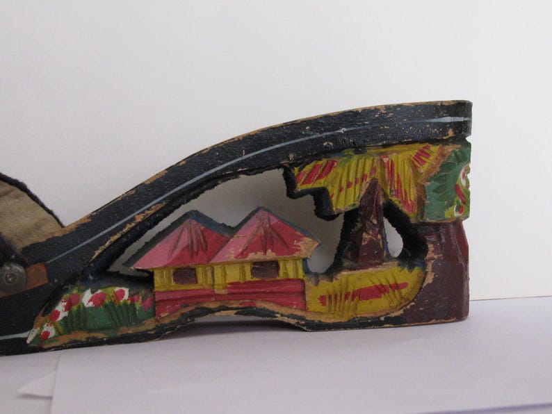 Peut inclure: Un talon de chaussure en bois noir pr&eacute;sentant une d&eacute;coupe peinte &agrave; la main repr&eacute;sentant deux petites maisons aux toits rouges et aux murs jaunes, entour&eacute;es de palmiers verts et de fleurs rouges. La peinture est us&eacute;e et &eacute;caill&eacute;e.