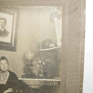 May include: A vintage black and white photograph of a woman seated in a room. The woman is wearing a dark dress with a white lace collar. A framed portrait hangs on the wall, and a table with a plant is visible.