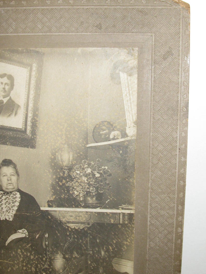 May include: A vintage black and white photograph of a woman seated in a room. The woman is wearing a dark dress with a white lace collar. A framed portrait hangs on the wall, and a table with a plant is visible.