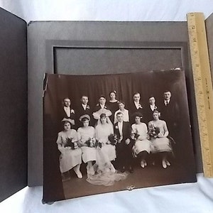 May include: A vintage sepia-toned photograph of a wedding party, featuring a bride, groom, and several attendants. The photo is in a dark gray album with a wooden ruler for scale. The image is slightly damaged.