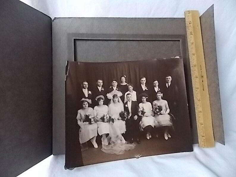 May include: A vintage sepia-toned photograph of a wedding party, featuring a bride, groom, and several attendants. The photo is in a dark gray album with a wooden ruler for scale. The image is slightly damaged.