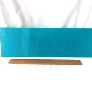 May include: A rectangular, turquoise sign with a hole in the upper left corner. A wooden ruler, approximately 30 cm long, rests below the sign. The background is white.
