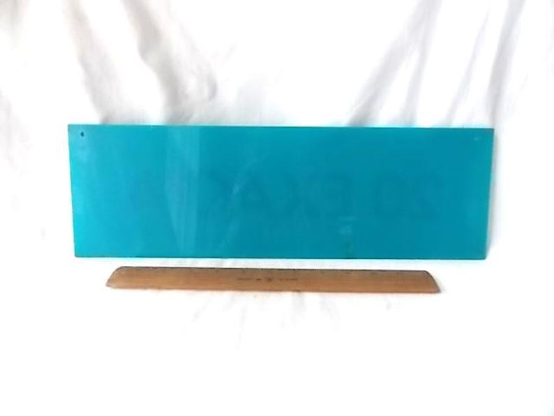 May include: A rectangular, turquoise sign with a hole in the upper left corner. A wooden ruler, approximately 30 cm long, rests below the sign. The background is white.