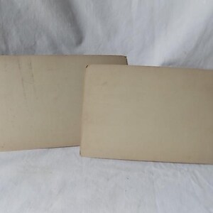 May include: Two rectangular, off-white cards are slightly overlapping, with rounded corners. The cards have a slightly aged appearance, with some minor discoloration and wear visible on their surfaces. The background is a white, textured fabric.