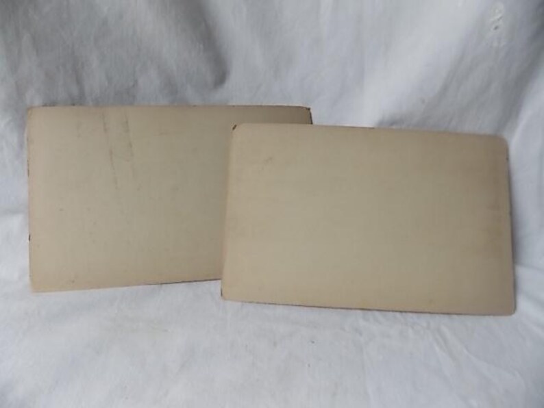 May include: Two rectangular, off-white cards are slightly overlapping, with rounded corners. The cards have a slightly aged appearance, with some minor discoloration and wear visible on their surfaces. The background is a white, textured fabric.