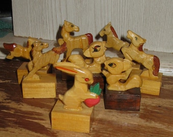May include: A collection of seven small, hand-carved wooden animal figurines mounted on square wooden bases. The set includes horses, a squirrel, and a rabbit holding a red and green carrot. Each figure features natural wood tones with painted details.