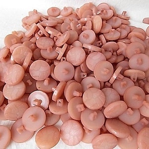 May include: A pile of light peach-colored buttons. The buttons are round with a slightly textured surface and a four-hole shank on the back. Some buttons have a decorative pattern on the front.