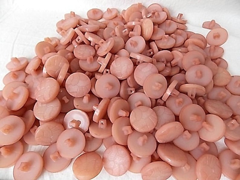 May include: A pile of light peach-colored buttons. The buttons are round with a slightly textured surface and a four-hole shank on the back. Some buttons have a decorative pattern on the front.