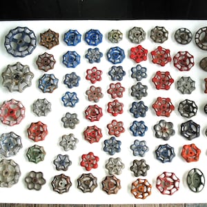 May include: A collection of various metal valve handles arranged in a grid. The handles are round, star-shaped, or oval, featuring colors including red, blue, grey, and rusted brown. Each handle has a central opening for attachment to a valve stem.