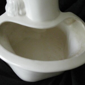May include: A white ceramic planter with a unique, curved design. The planter has a wide, open bowl shape with a rounded rim and a small decorative element at the top. The interior is a light beige color.