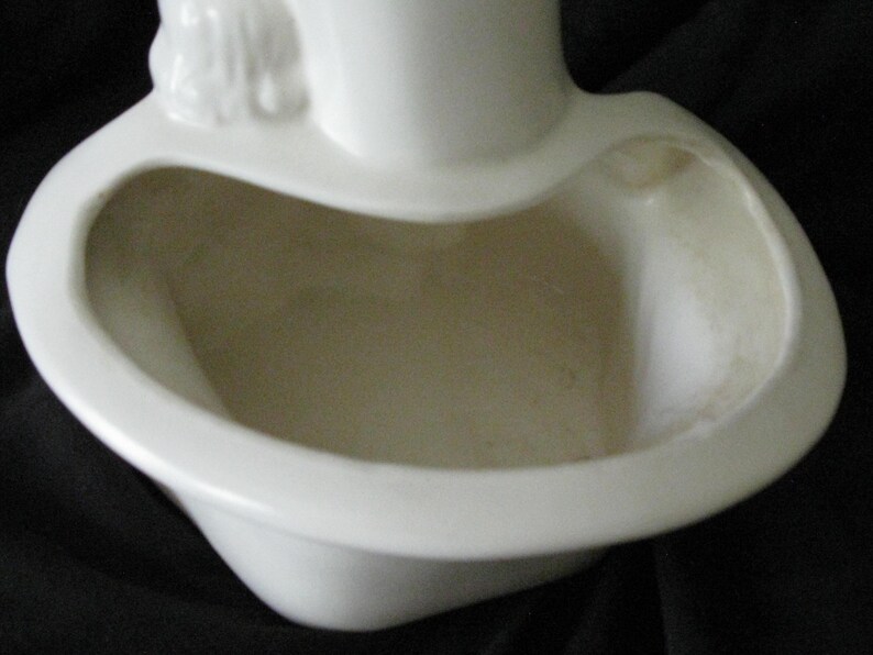 May include: A white ceramic planter with a unique, curved design. The planter has a wide, open bowl shape with a rounded rim and a small decorative element at the top. The interior is a light beige color.