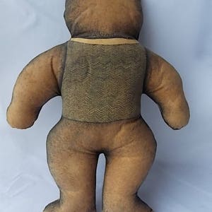 May include: A vintage teddy bear plush toy, viewed from the back. The bear has a brown body with darker brown accents on the head and limbs. It wears a vest with a zigzag pattern and a yellow collar.