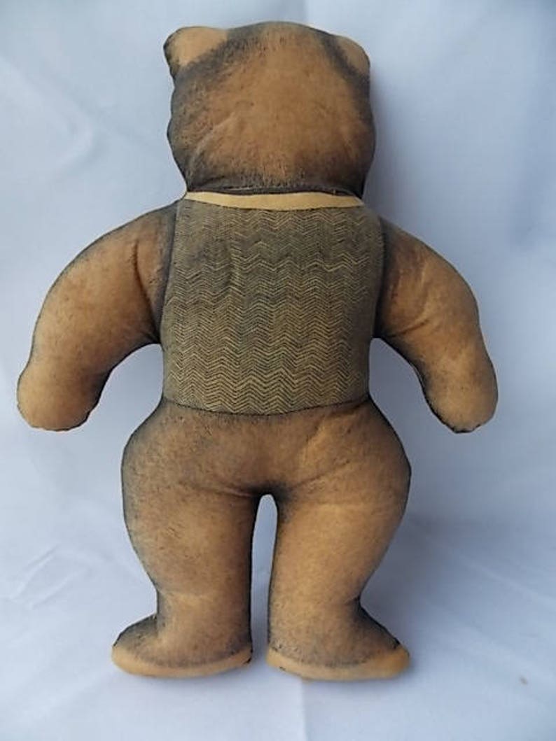 May include: A vintage teddy bear plush toy, viewed from the back. The bear has a brown body with darker brown accents on the head and limbs. It wears a vest with a zigzag pattern and a yellow collar.