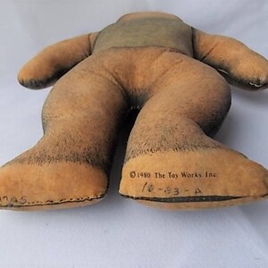 May include: A vintage plush toy in the shape of a bear, lying on its back. The bear is primarily tan with darker brown accents on the feet and legs. The text "&copy;1980 The Toy Works Inc" is printed on the foot.