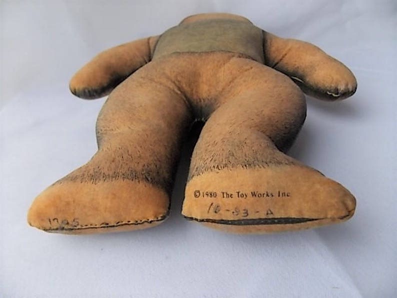 May include: A vintage plush toy in the shape of a bear, lying on its back. The bear is primarily tan with darker brown accents on the feet and legs. The text "&copy;1980 The Toy Works Inc" is printed on the foot.