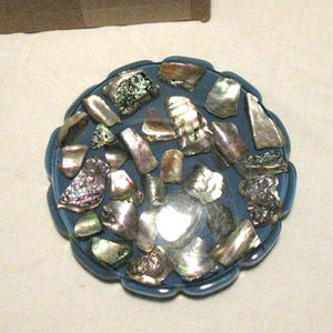 May include: A blue, scalloped-edge dish filled with iridescent abalone shell fragments. The shells are various sizes and shapes, showing silver, green, and purple hues. A decorative item.