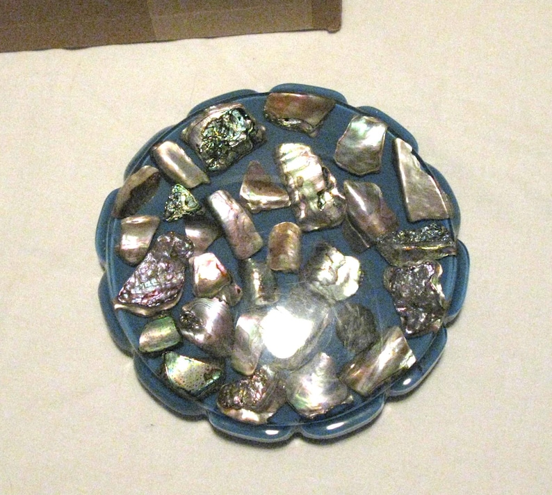 May include: A blue, scalloped-edge dish filled with iridescent abalone shell fragments. The shells are various sizes and shapes, showing silver, green, and purple hues. A decorative item.