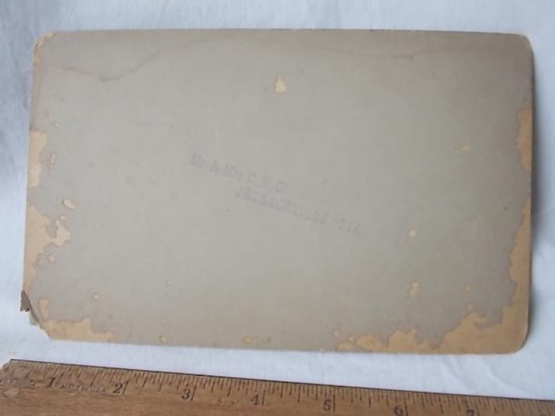 May include: A vintage photograph with a light tan background. The edges show signs of wear and discoloration. Faint text is visible, reading "Mr. & Mrs. C. Rigby, Jacksonville, Fla.". A wooden ruler is at the bottom.