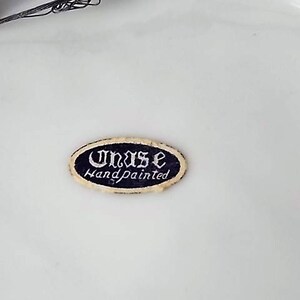 Peut inclure: Un autocollant ovale avec le texte "Unas Handpainted" en &eacute;criture blanche sur un fond bleu fonc&eacute;. L'autocollant a une bordure dor&eacute;e et est pos&eacute; sur un fond blanc.