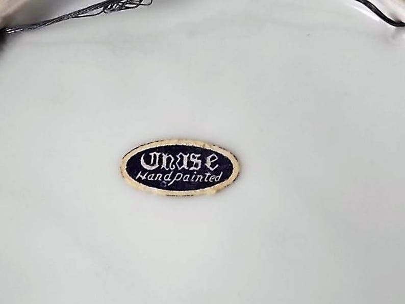 Peut inclure: Un autocollant ovale avec le texte "Unas Handpainted" en &eacute;criture blanche sur un fond bleu fonc&eacute;. L'autocollant a une bordure dor&eacute;e et est pos&eacute; sur un fond blanc.
