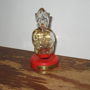May include: A vintage perfume bottle in the shape of a seated figure with a clear glass body and gold accents. The figure sits atop a red base with the text "DEVON VIOLETS" in gold lettering. The bottle is displayed on a wooden surface.
