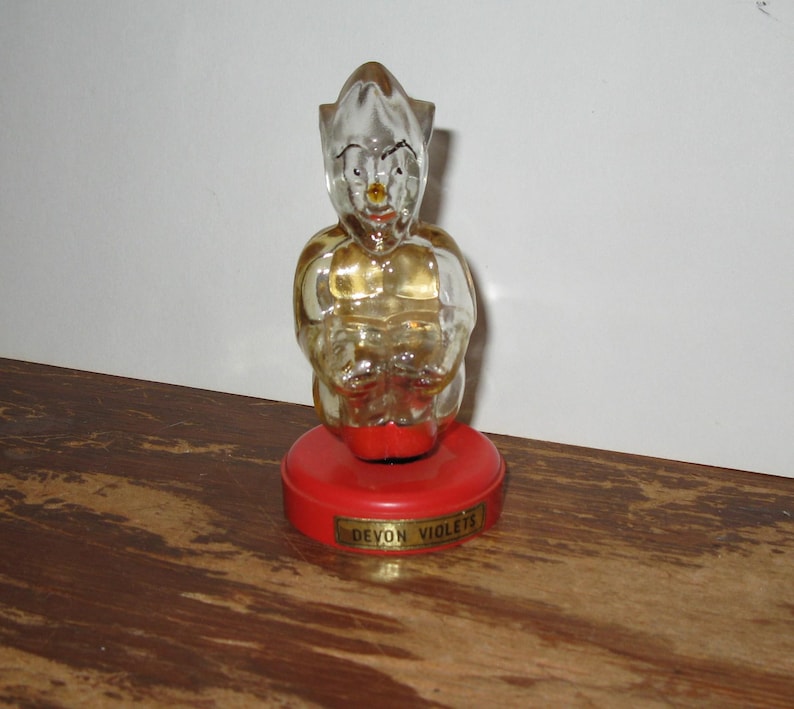 K&ouml;nnte beinhalten: Vintage Parf&uuml;mflasche in Form einer sitzenden Figur mit klarem Glask&ouml;rper und goldenen Akzenten. Die Figur steht auf einem roten Sockel mit dem Text "DEVON VIOLETS" in goldener Schrift. Die Flasche steht auf einer Holzoberfl&auml;che.