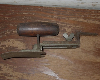 May include: A vintage metal tool featuring a dark brown wooden handle attached to a horizontal frame. The mechanism includes a metal wing nut for adjustment and a flat, notched metal plate extending from the base, showing signs of age and wear.